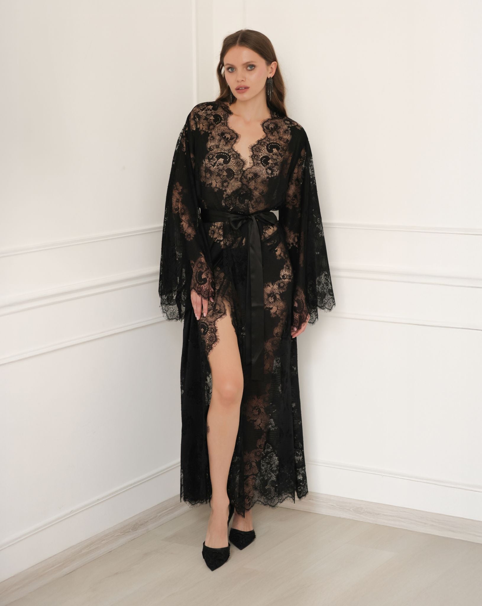 Full-length pose against a white wall in a black lace gown, showcasing a high leg slit and intricate fabric texture.