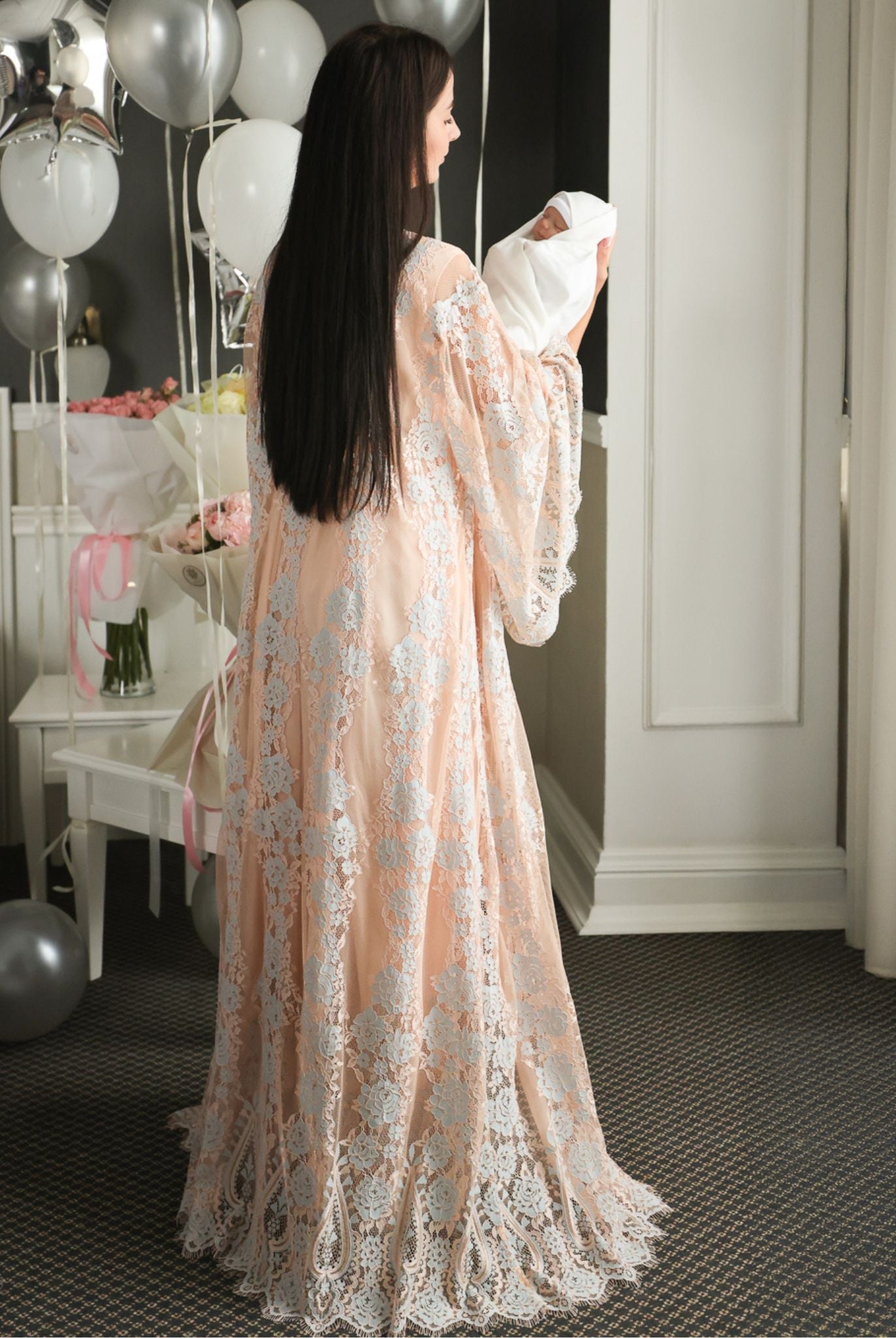 Full-length rear view of a mother holding her baby, showcasing the graceful drape of the long lace maternity robe.