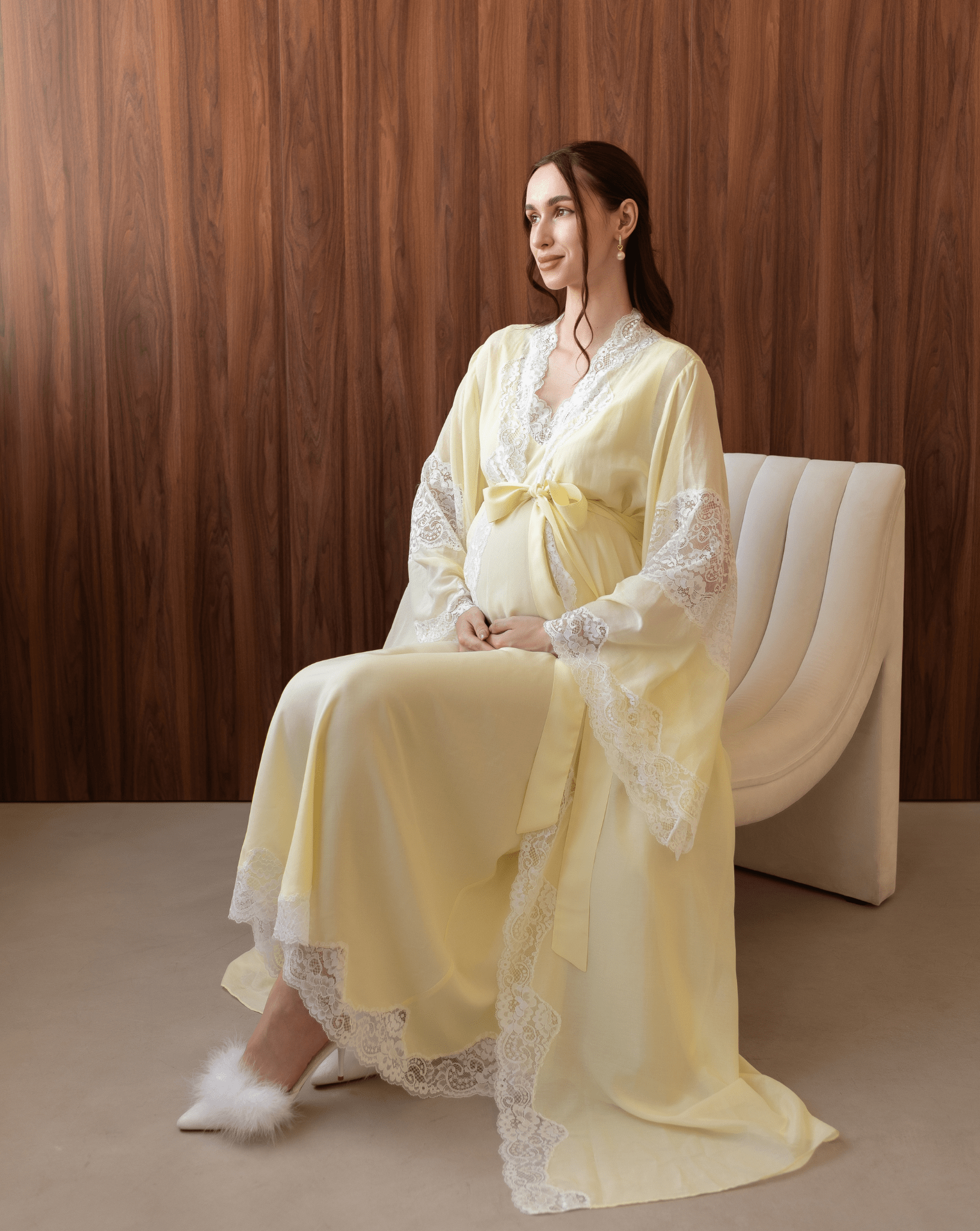 Pregnant woman sitting on a white chair, gently holding her belly in a comfortable yellow cotton lace ensemble.