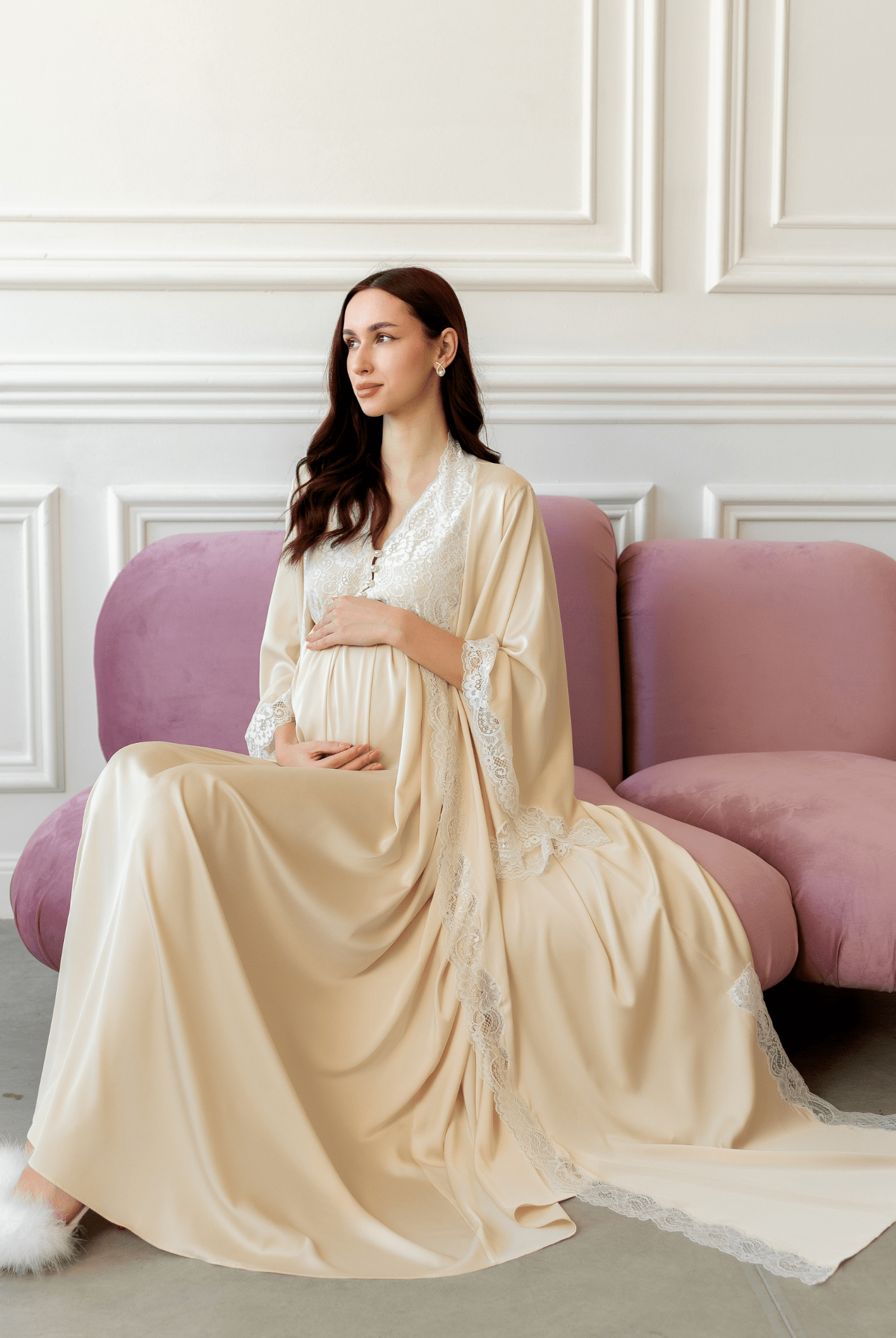 Sitting on a purple sofa in a vanilla robe, gently embracing a baby bump in a cozy and serene setting.