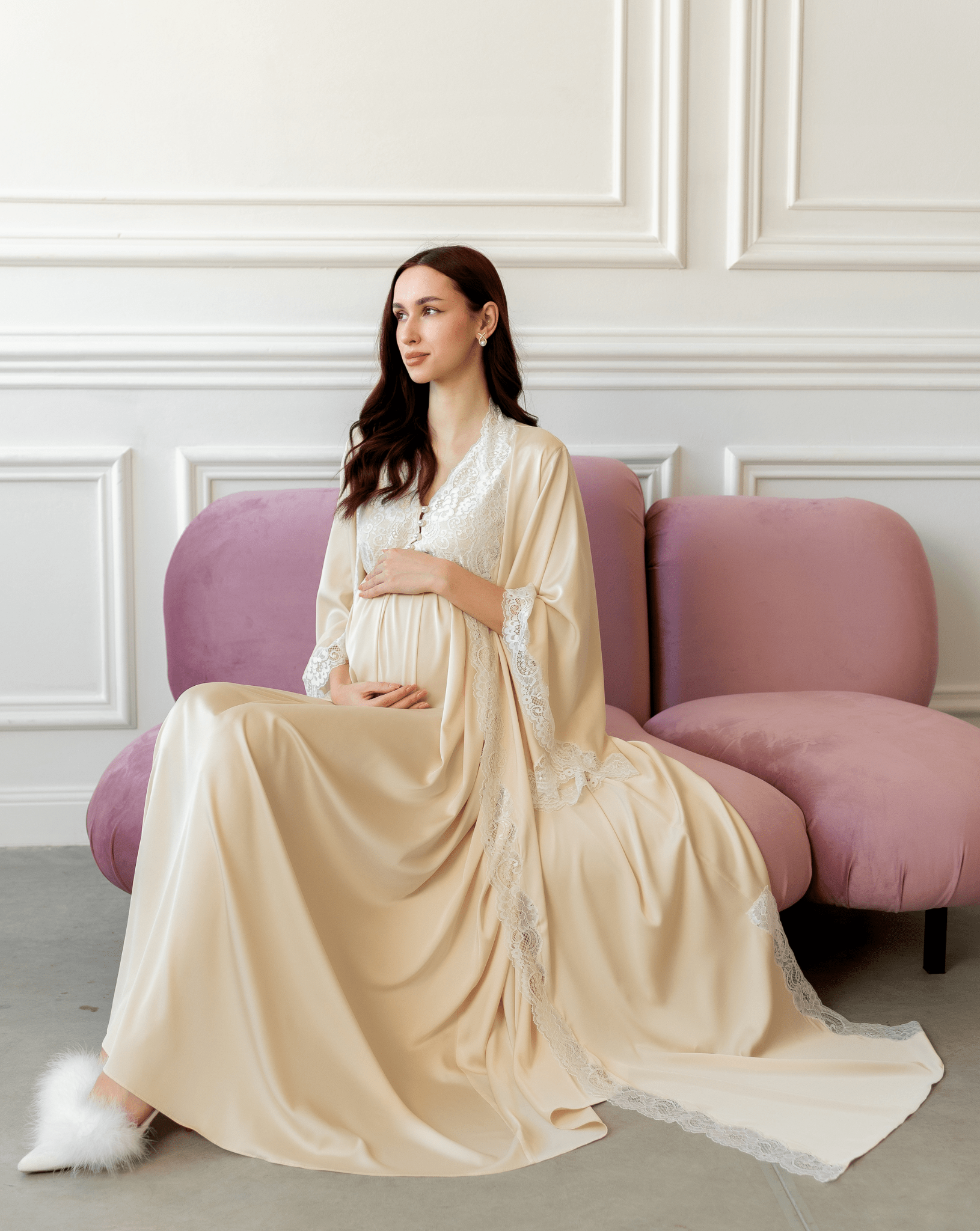 Sitting on a purple sofa in a vanilla robe, gently embracing a baby bump in a cozy and serene setting.