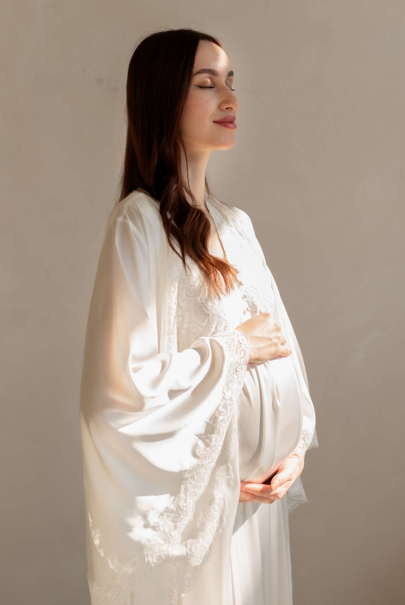 Gentle waist-up shot in a white set with sunlight, softly embracing a baby bump for a tender look.