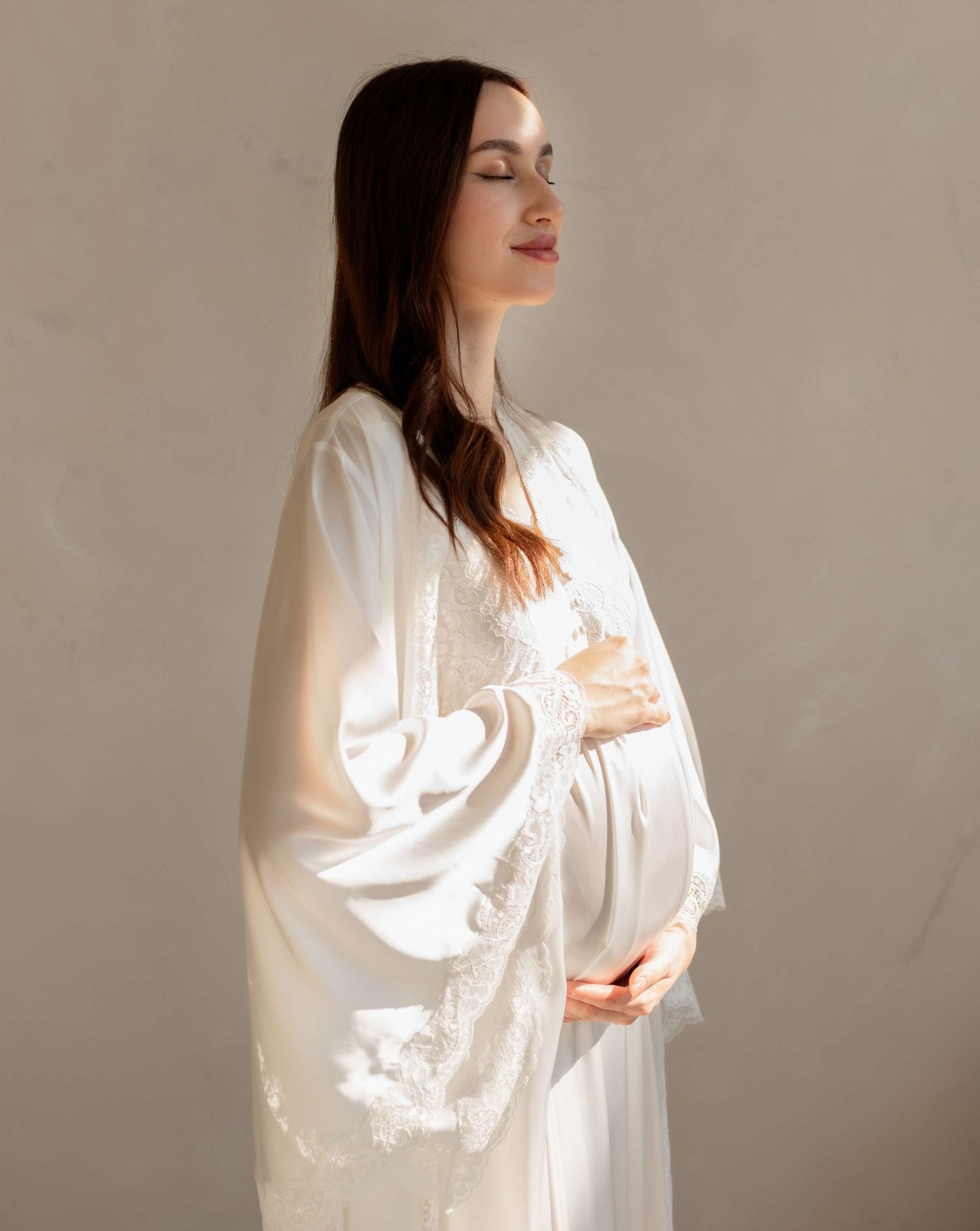 Gentle waist-up shot in a white set with sunlight, softly embracing a baby bump for a tender look.