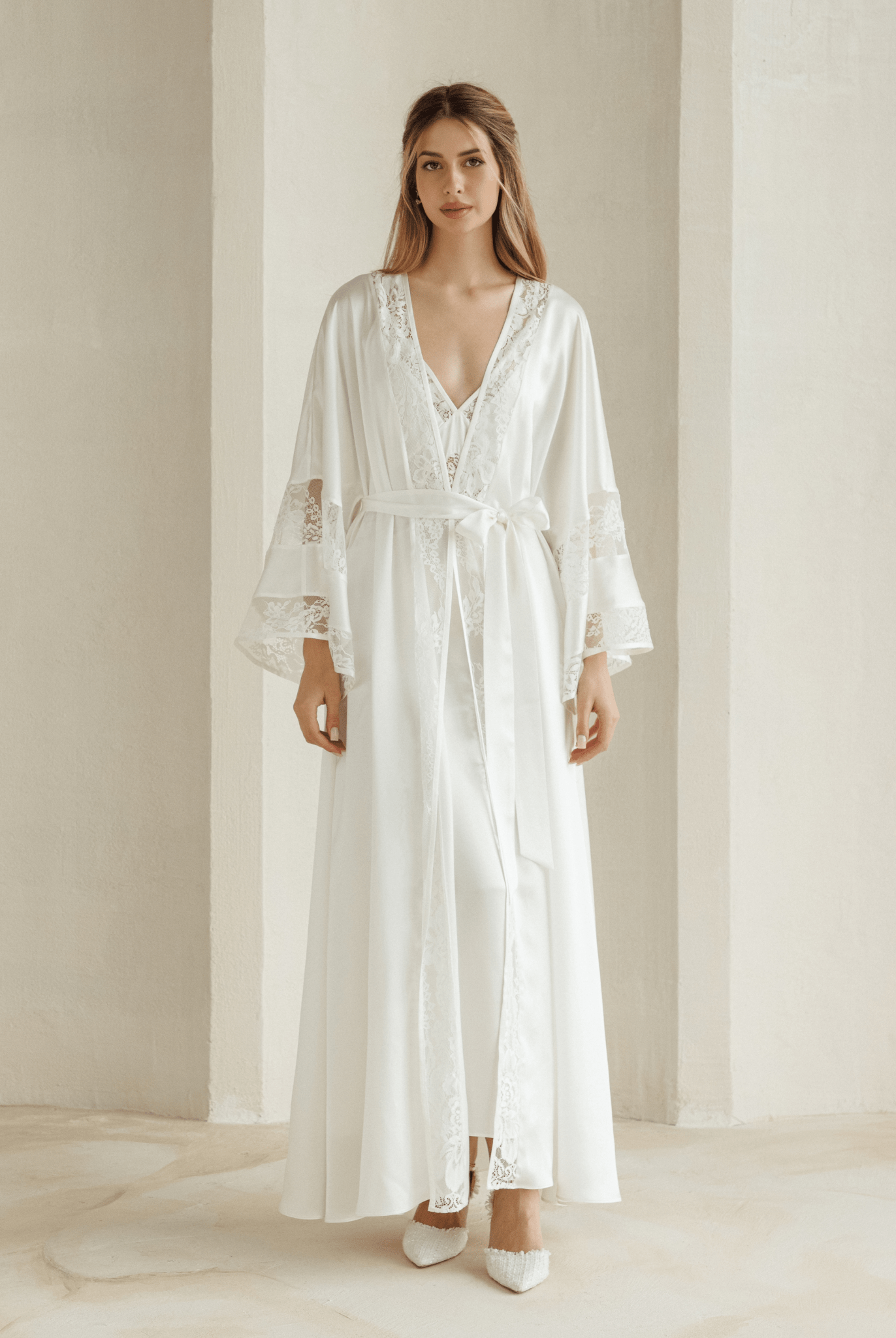 Woman wearing white silk robe over matching lace nightgown standing in soft natural light