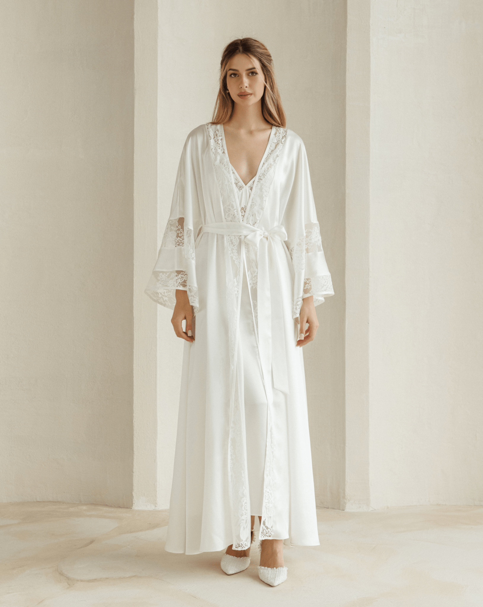 Woman wearing white silk robe over matching lace nightgown standing in soft natural light