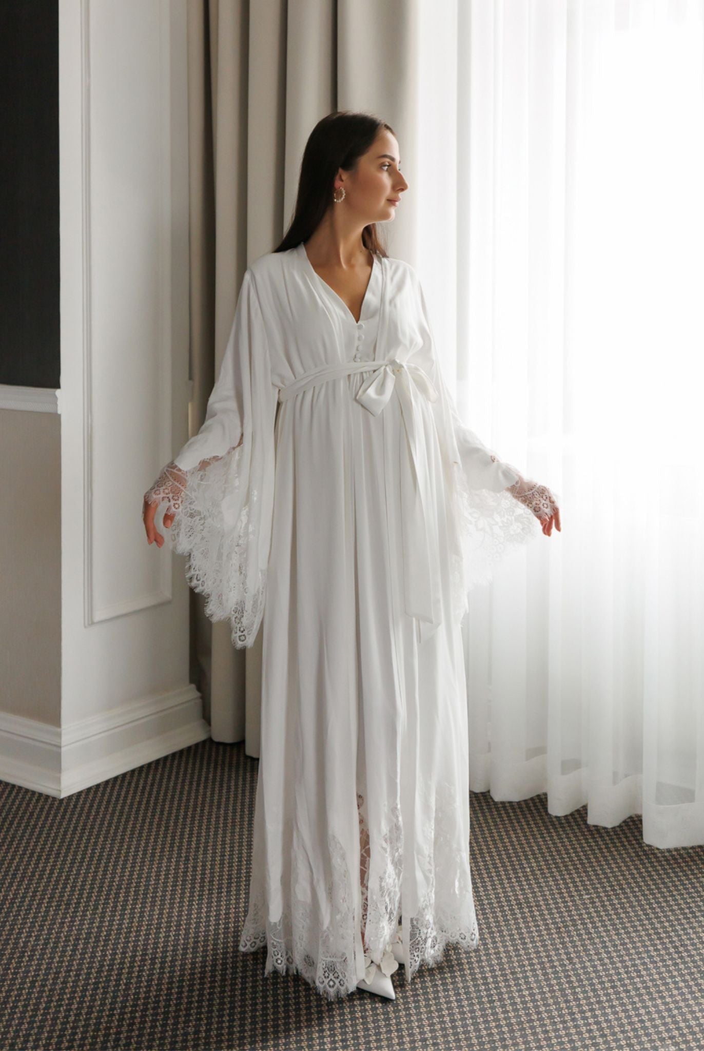 Standing by a window in a white ensemble, showcasing the wide sleeves decorated with delicate lace at the ends.
