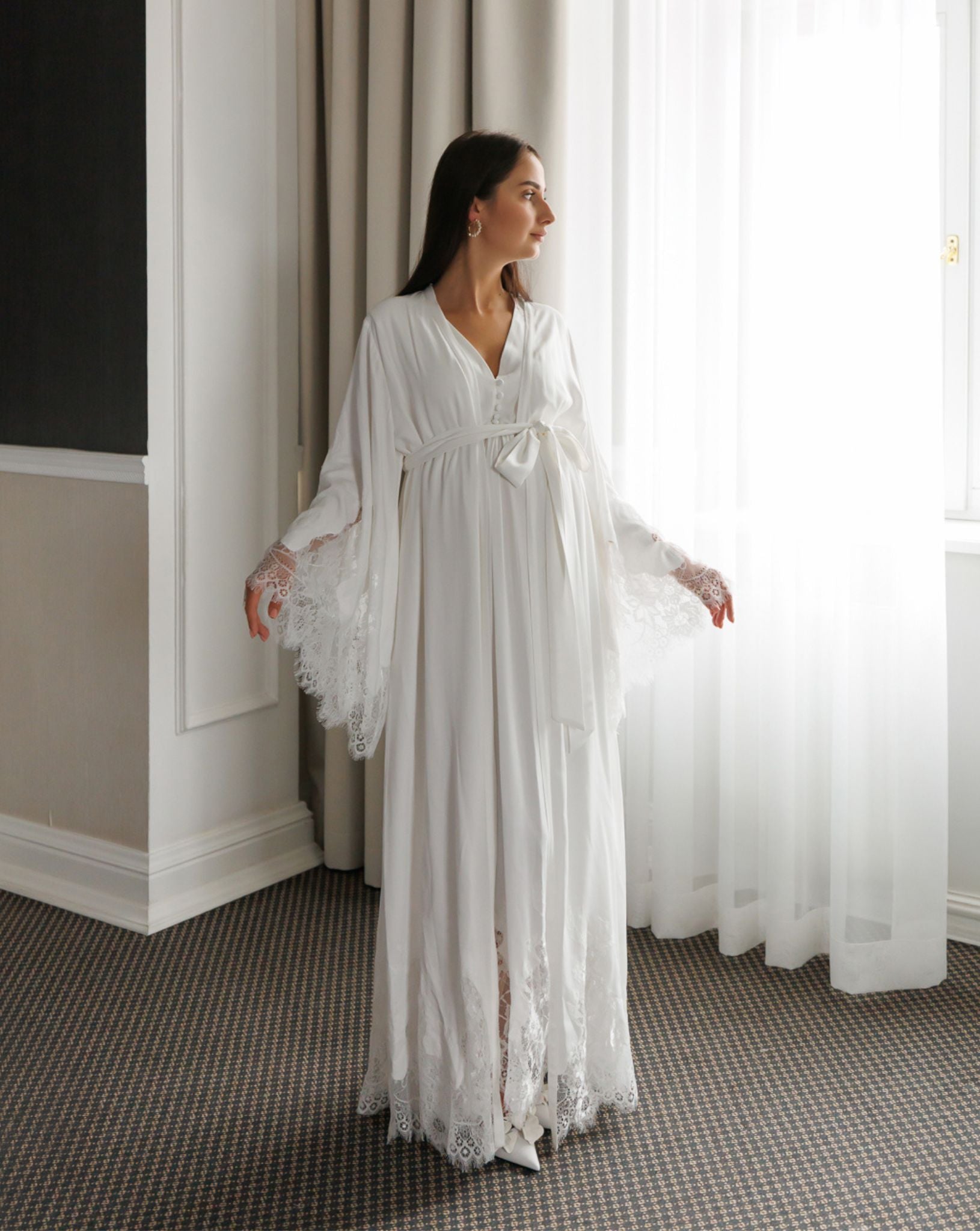 Standing by a window in a white ensemble, showcasing the wide sleeves decorated with delicate lace at the ends.