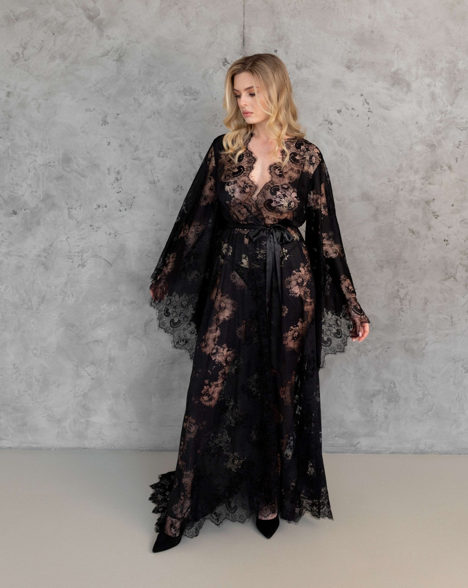 Model in sheer black lace robe with flared sleeves, posing in front of textured grey wall – KÂfemme