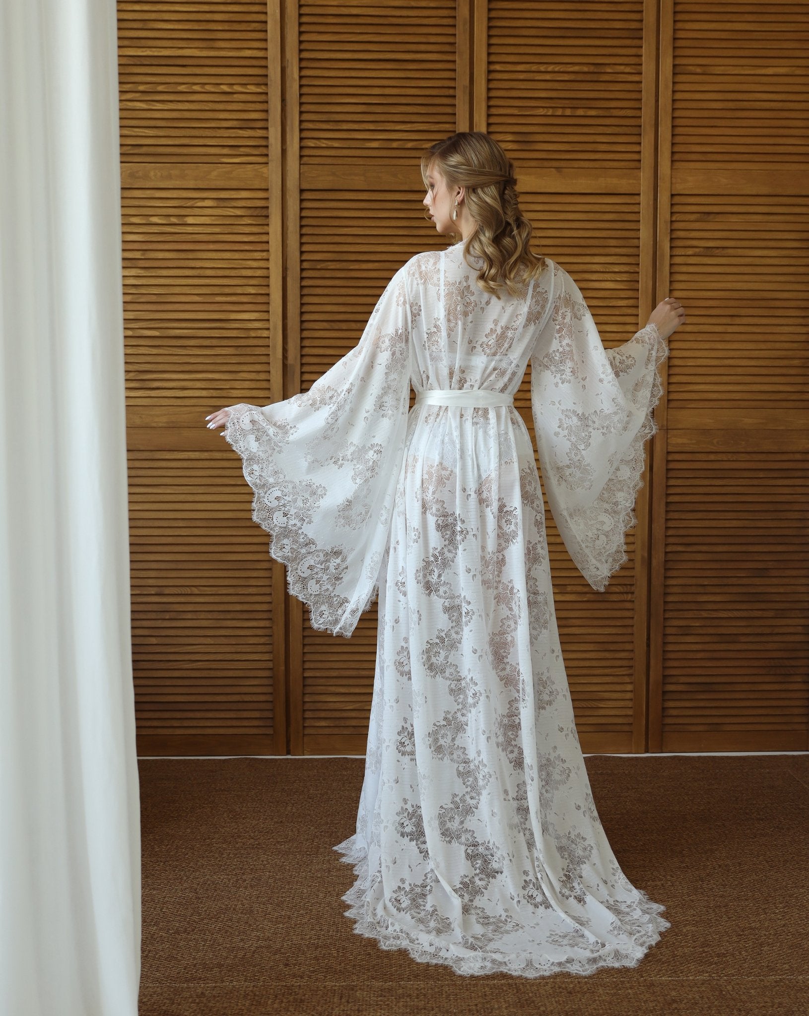 Back view of white lace robe with sweeping train, posed by wooden backdrop – KÂfemme