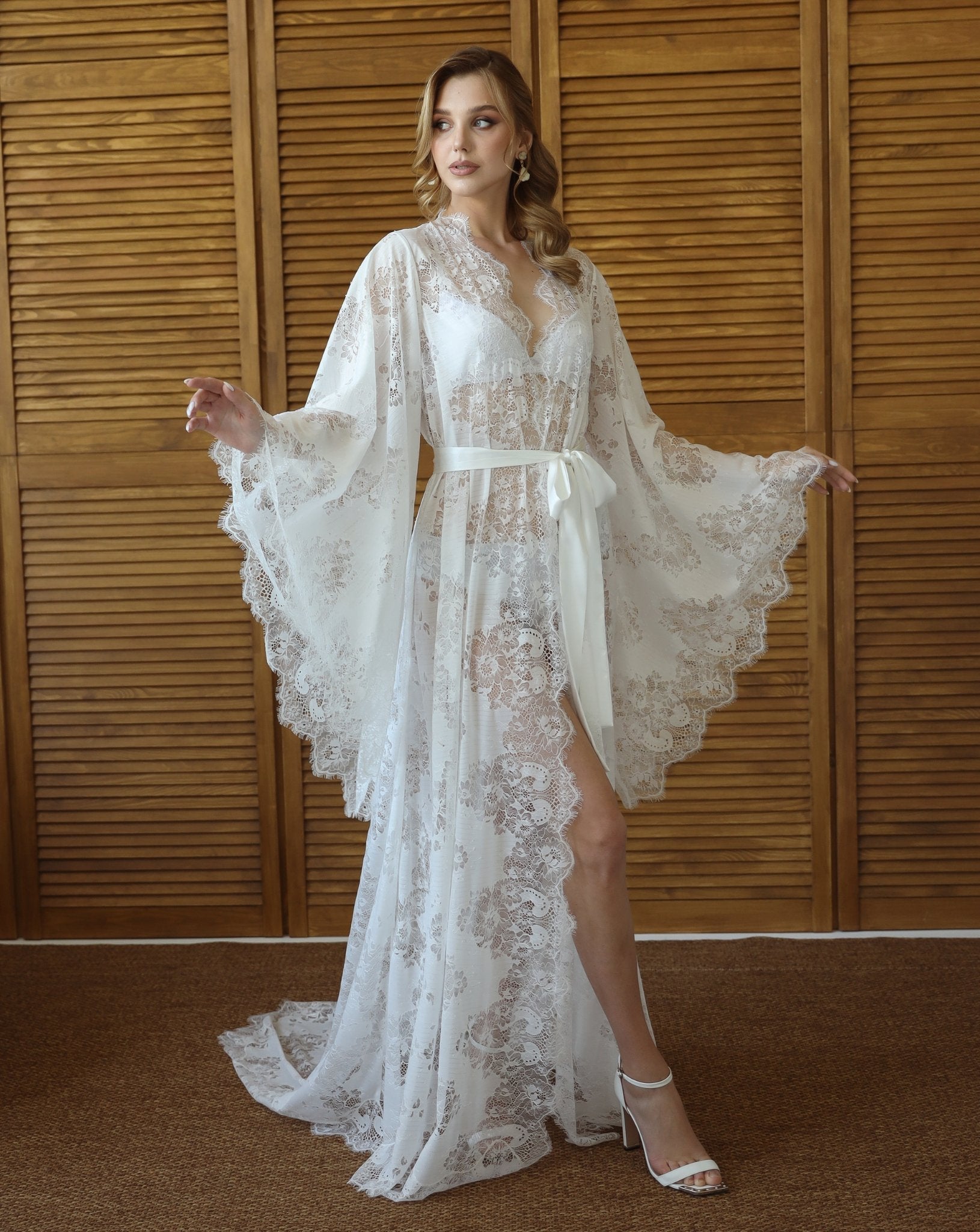 Woman in long white lace bridal robe with flared sleeves, standing by wooden panels – KÂfemme