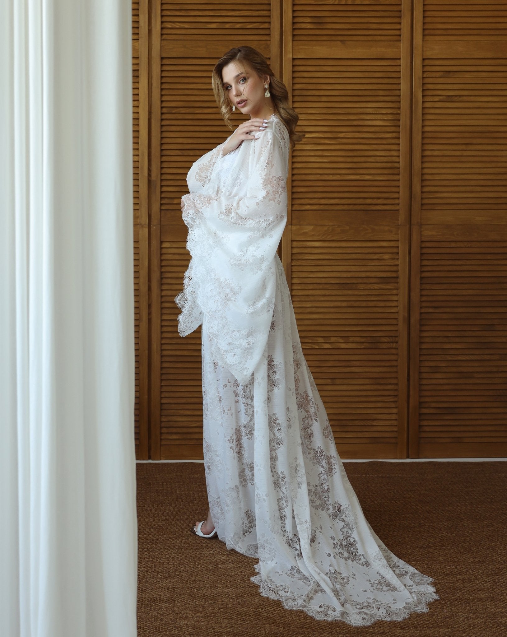 Model in sheer white lace robe with flared sleeves, standing against wooden background – KÂfemme