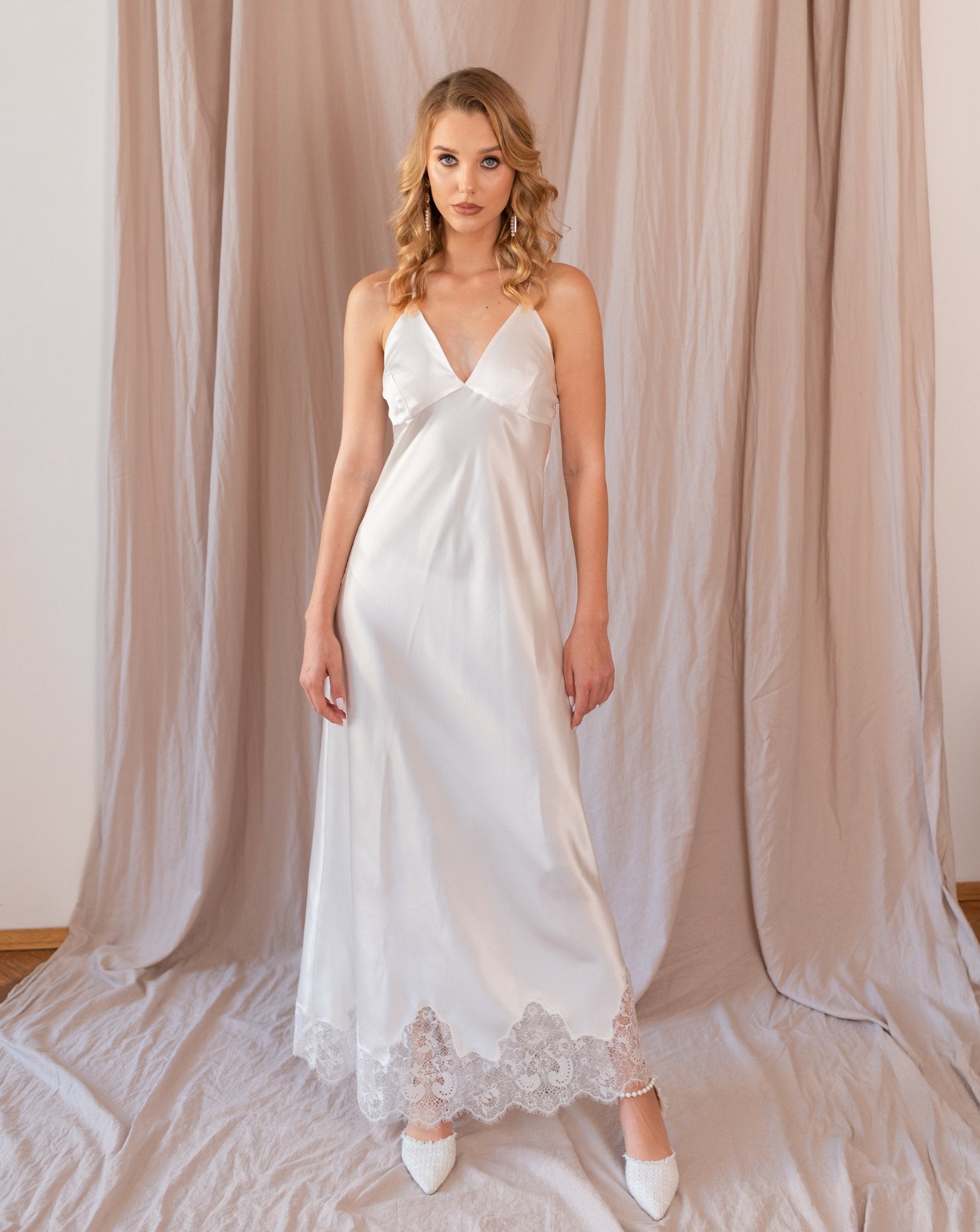 Woman standing in satin white nightgown with lace trim, light pink curtain background