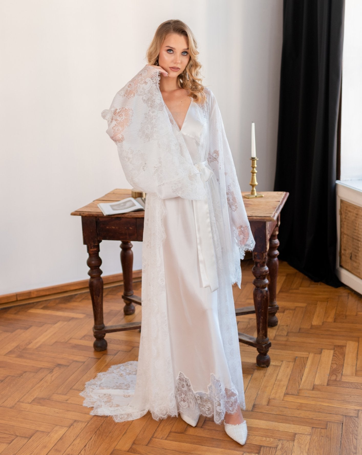 Woman standing in a white lace bridal robe and silk nightgown in a wood-floored room