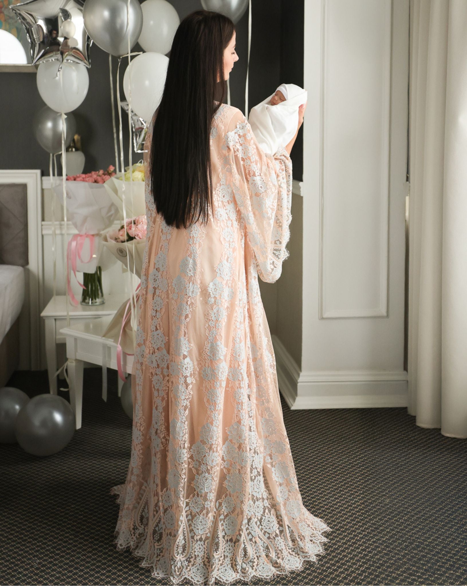 Full-length rear view of a mother holding her baby, showcasing the graceful drape of the long lace maternity robe.