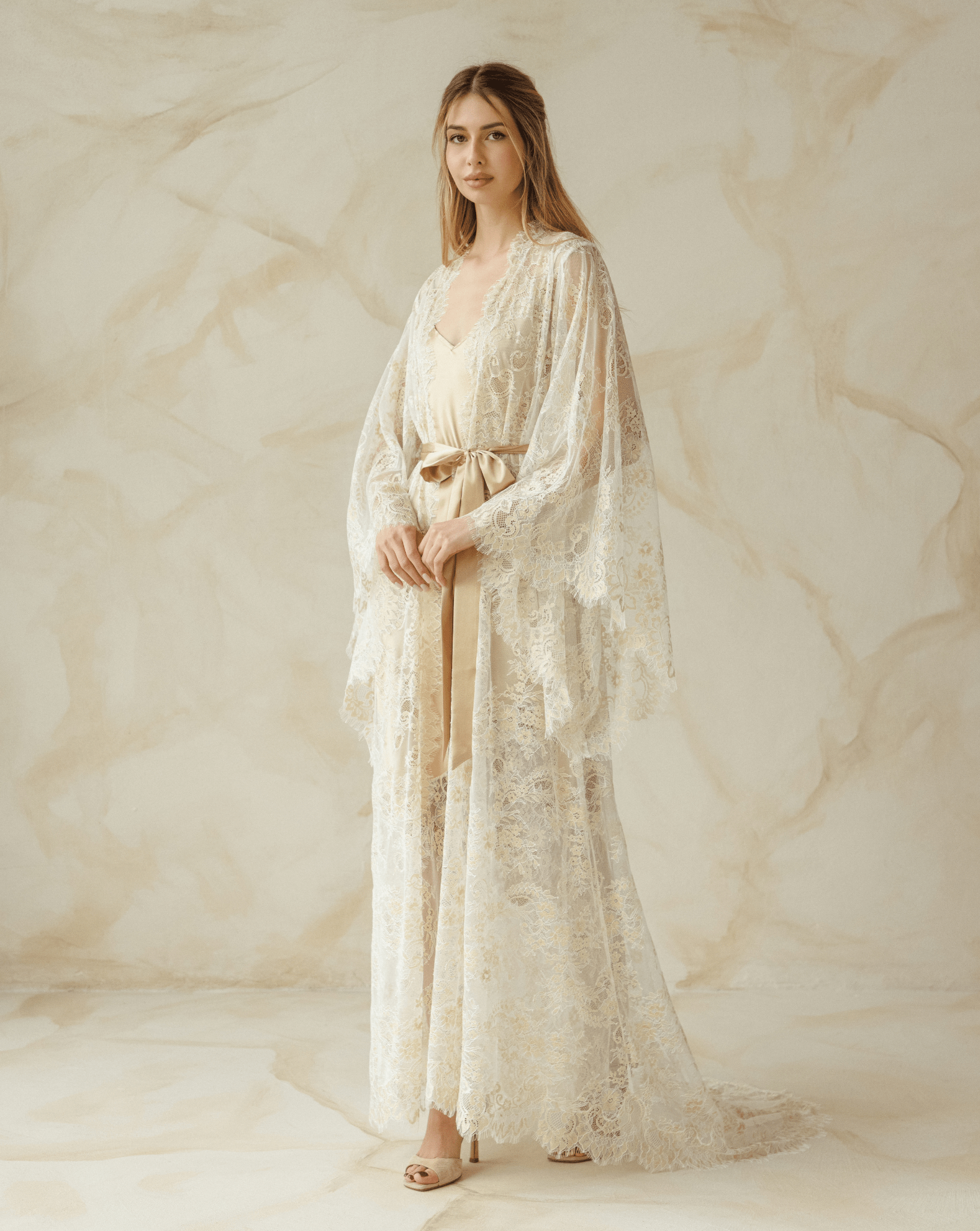 Side view of model gently draping long lace robe in soft beige tones