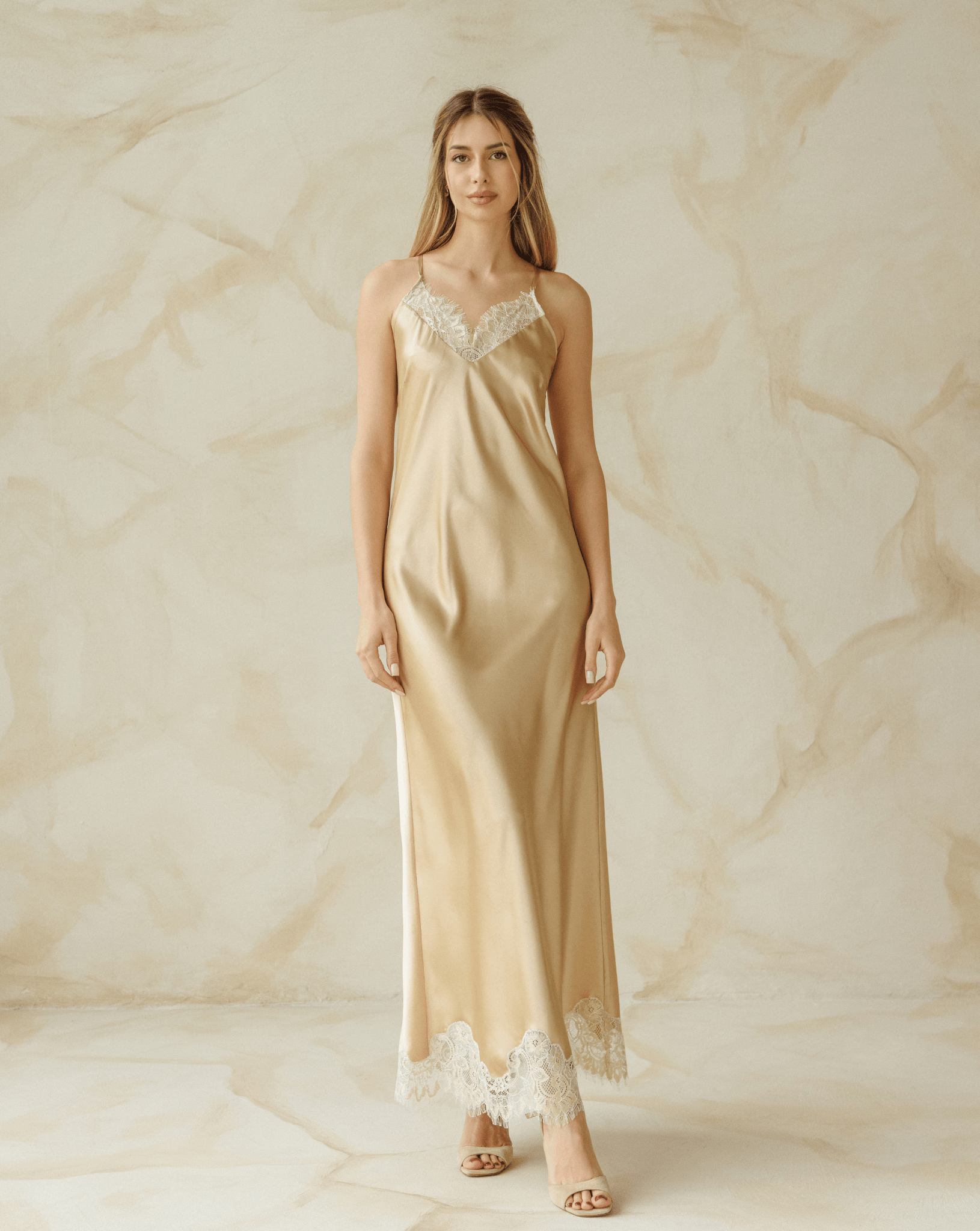 Full-length view of a woman in a champagne silk nightgown standing against marble wall